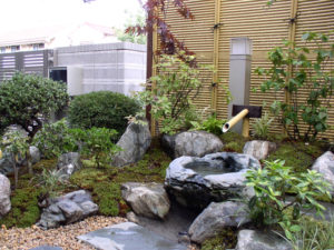 自然石のつくばいを庭の中心にし、背景には人工竹垣で目隠ししています